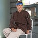A man sitting on a stool wearing an Uncommon Chef Orleans brown long sleeve chef coat.