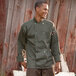 A man in a Uncommon Chef Orleans olive long sleeve chef coat standing next to a wooden fence.