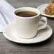 A Tuxton eggshell china saucer with a cup of coffee and a pastry on a plate.