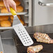 A person using a Mercer Culinary perforated turner with a yellow handle to cook chicken on a grill.