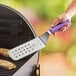 A hand using a Mercer Culinary purple perforated turner to cut food on a grill.