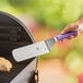 A hand holding a Mercer Culinary Millennia turner with a purple handle over a grill.