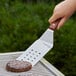 A person using a Dexter-Russell perforated turner to flip a burger on a grill.