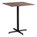 A square bar height table with a textured walnut finish top and a black cross base plate.