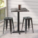A Lancaster Table & Seating black metal square counter height table with a cross base plate and two black stools on a porch.