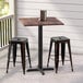 A Lancaster Table & Seating square counter height table with a textured walnut finish and cross base plate with two black stools on an outdoor patio.