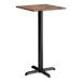 A square bar height table with a textured oak finish top and a black cross base plate.