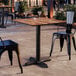 A square standard height table with a textured Yukon oak finish and a black cross base plate, shown with black metal chairs in an outdoor setting.