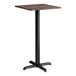 A square bar height table with a textured walnut finish top and a black cross base plate.