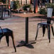 A square standard height table with a textured Yukon oak finish and a black cross base plate, shown with two black metal chairs in an outdoor setting.