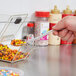 A hand holding a clear topping dispenser spoon filled with colorful candy over a container of candy.