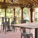 A Lancaster Table & Seating Excalibur bar height table with a textured mixed plank finish and cross base plate on a covered patio with chairs.