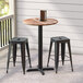 A Lancaster Table & Seating round counter height table with a textured wood finish and cross base plate on a patio with two black stools.