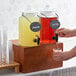 A person pouring lemonade from an Acopa glass beverage dispenser with a black label on a counter.