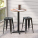 A Lancaster Table & Seating Excalibur counter height table with a textured plank finish and two black bar stools on a porch.