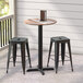 A Lancaster Table and Seating counter height table with a textured mixed plank finish and cross base plate on a porch with black bar stools.