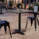 A square standard height table with a smooth Paladina finish and a cross base plate, set outdoors with two black metal chairs.