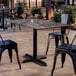 A rectangular standard height table with a smooth dark finish and a cross base plate, surrounded by metal chairs in an outdoor setting.