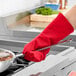 A person wearing a Tucker Safety red oven mitt holding a pan of food over a stove.