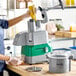 A commercial combination food processor with a gray and green base, a plastic bowl, and continuous feed attachment, shown in use on a kitchen counter.