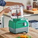 A green and gray AvaMix Revolution food processor with a clear batch bowl containing chopped vegetables.