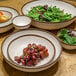 A white bowl with brown rim on a table with fruit and salad.