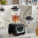 A commercial blender with a keypad control base and two large clear containers, one filled with iced liquid and the other empty, both with handles and lids.