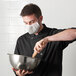 A man wearing a Mercer Culinary white face mask mixing food in a bowl.