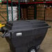 A black Rubbermaid tilt truck with wheels and a hinged dome lid in a warehouse.