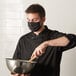 A man wearing a Mercer Culinary black face mask and holding a bowl.