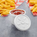 A white plastic container of ketchup with a Dart white plastic vented lid on a table with french fries.