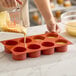 A red silicone baking mold with eight cylindrical compartments being filled with batter.