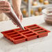 A red silicone baking mold with 12 rectangular compartments being filled with batter from a piping bag.