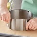 A person using an Ateco stainless steel adjustable round cake ring on a table in a professional kitchen.