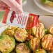 A hand holding a Lee Kum Kee Sriracha chili sauce packet over cooked brussels sprouts.