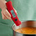 A person holding a Taylor red digital thermocouple thermometer over a pot of soup.