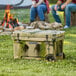 A CaterGator green and black camouflage cooler on grass with people sitting on a log.