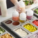 A person using a Choice stainless steel squeeze bottle holder to pour sauce over a tray of food.