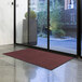 A Lavex solid burgundy entrance mat on the floor in front of a glass door.