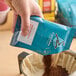 A hand holding a blue Caribou Coffee package pouring brown Caribou Coffee grounds into a coffee maker.