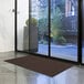 A brown Lavex Needle Rib entrance mat roll on the floor in front of a glass door.