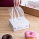 A hand using LK Packaging plastic deli wrap to bag a pink donut with sprinkles over a box of donuts.