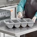 A woman in a blue apron holding a white Rubbermaid container with four sections of silverware in it.