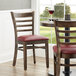 Two Lancaster Table & Seating wood ladder back chairs with burgundy vinyl seats next to a table in a restaurant dining area.