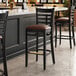 A Lancaster Table & Seating black wood ladder back bar stool with a dark brown vinyl seat on a black surface.