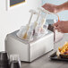 A person using a Steril-Sil stainless steel condiment dispenser to pour liquid into a cup on a counter.