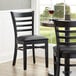 A Lancaster Table & Seating black wood ladder back chair with black vinyl cushion at a table in a restaurant.