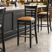 A Lancaster Table & Seating black wood bar stool with a light brown cushion on the seat.