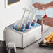 A person pouring ketchup into a Steril-Sil stainless steel condiment dispenser with blue pump lids.