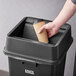 A hand holding a coffee cup in a Lavex black square trash can with a swing lid.